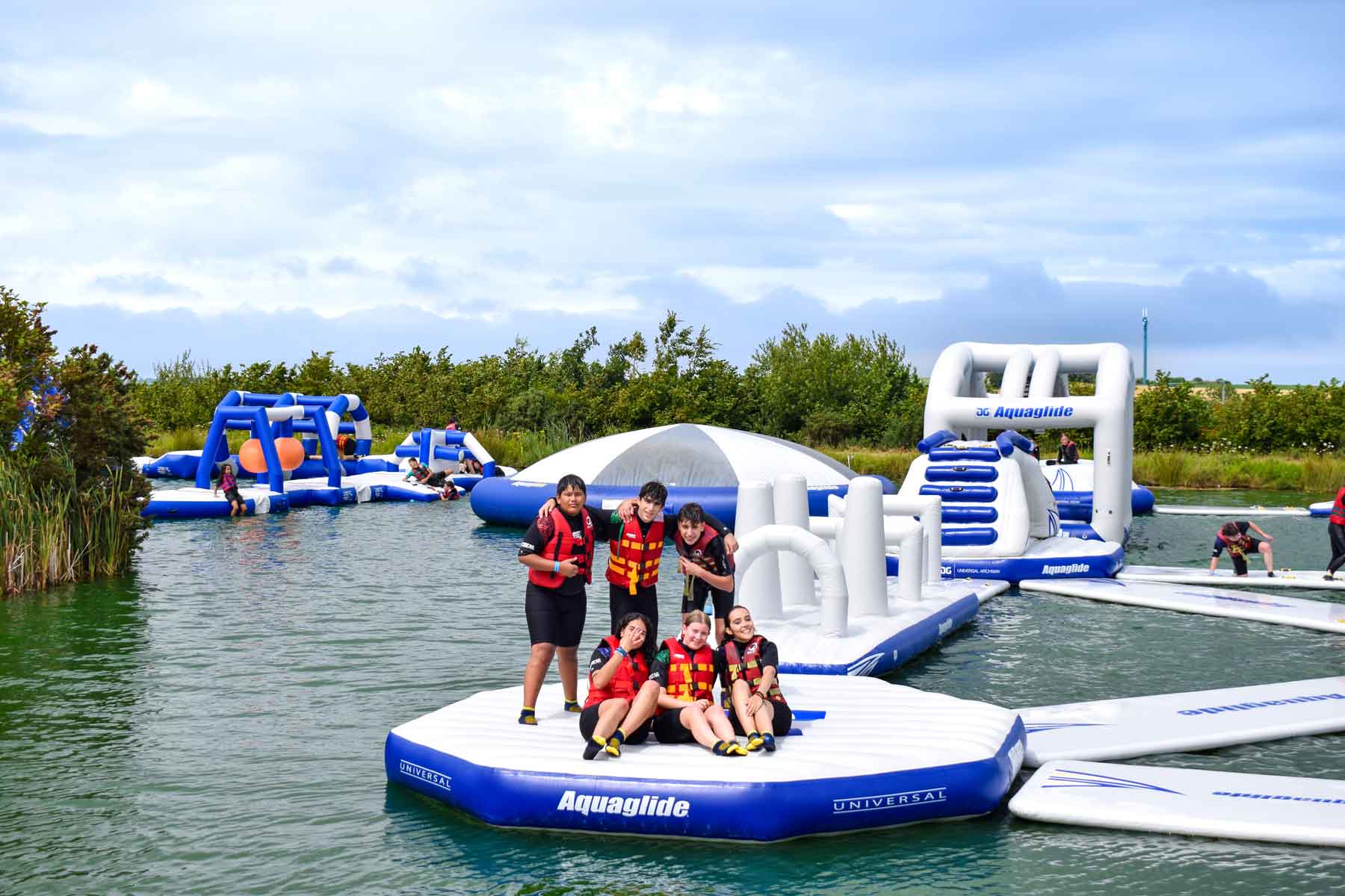 Camp students in buoyancy aids and helmets on an inflatable water park platform on a lake during an outdoor activity day