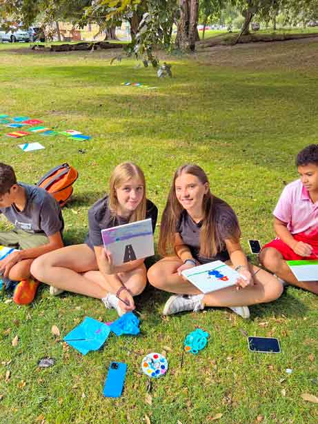 Camp students sitting on the grass on a hillside with notebooks and craft materials during a creative session