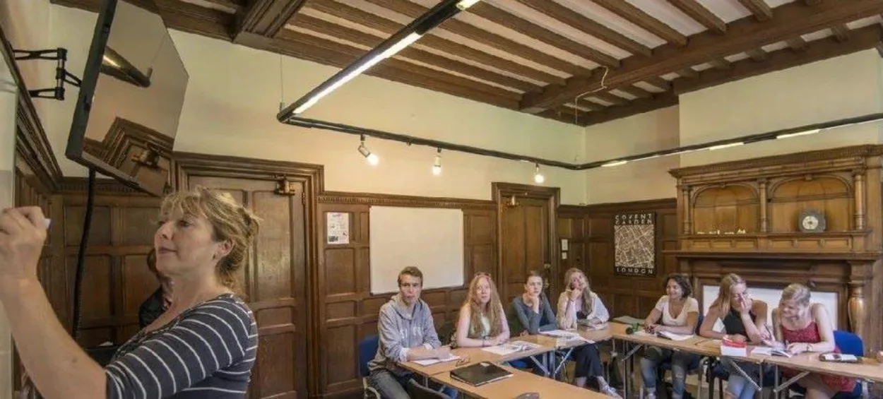 A group of teenage English students look towards the teacher as she writes on the board.
