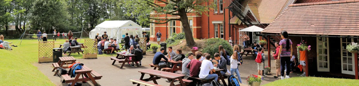 A photo of students sitting at picnic tables chatting together during a break in activities at Loxdale, which is the home to ELC Brighton's summer camp for teenagers. You can see the red brick building in the background and a white gazebo which has been erected on the lawn. The sun is shining.