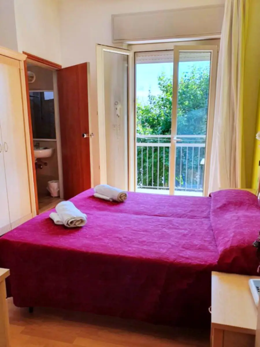 A hotel room with made-up beds, folded towels, an en-suite shower room and a balcony door open to greenery