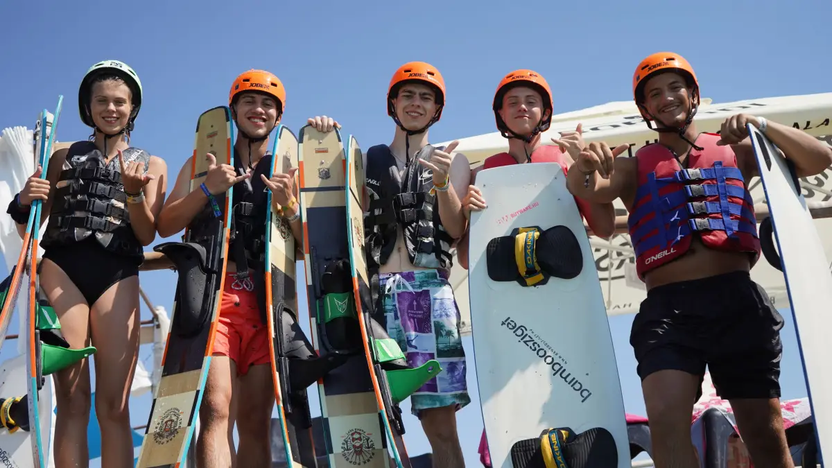Five participants in buoyancy vests and helmets holding wakeboards at the cable park