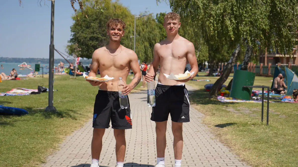 Two boys carrying plates of food along the beach path, with sunbathers and the lake behind