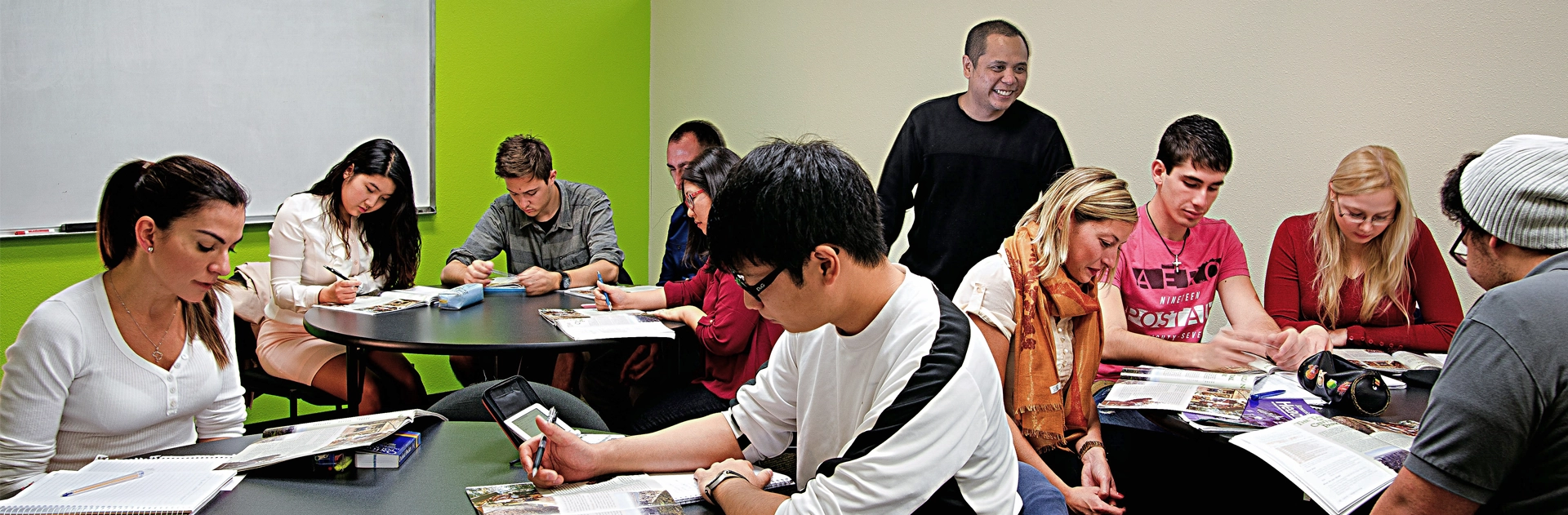 Students working with printed materials around a round table in a green-walled classroom at LSI San Diego, with a teacher standing behind them.