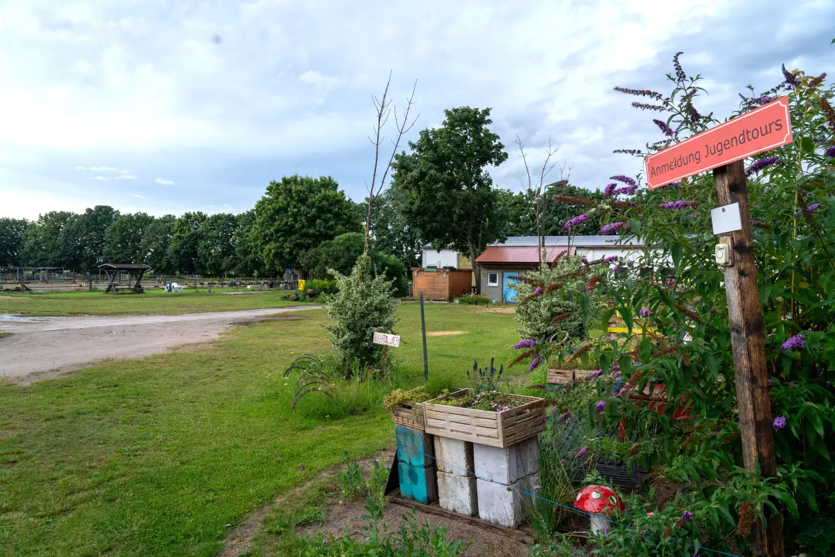 The entrance to the yard with a wooden signpost, flower beds and the sand riding ground behind