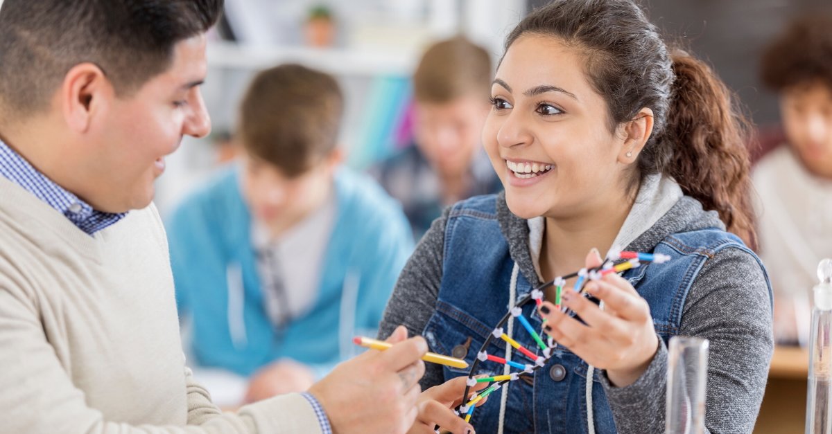 Student holding a plastic model of a DNA double helix and talking with an instructor in a classroom