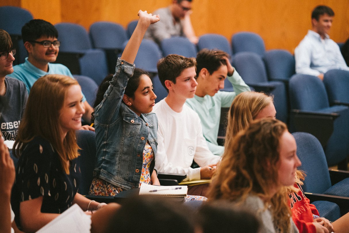 High school students seated in a tiered lecture hall, one raising her hand to answer
