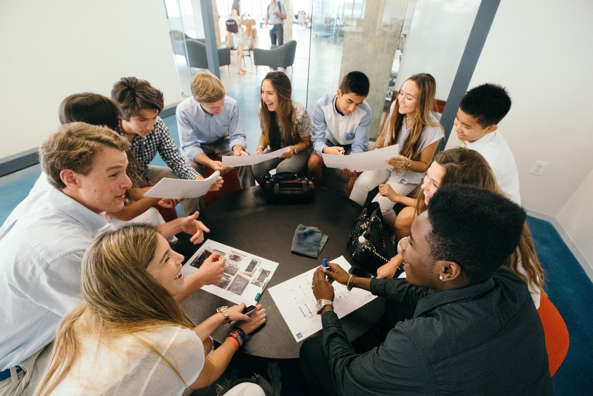 High school students sitting around a table with printed papers and laptops, working in a group