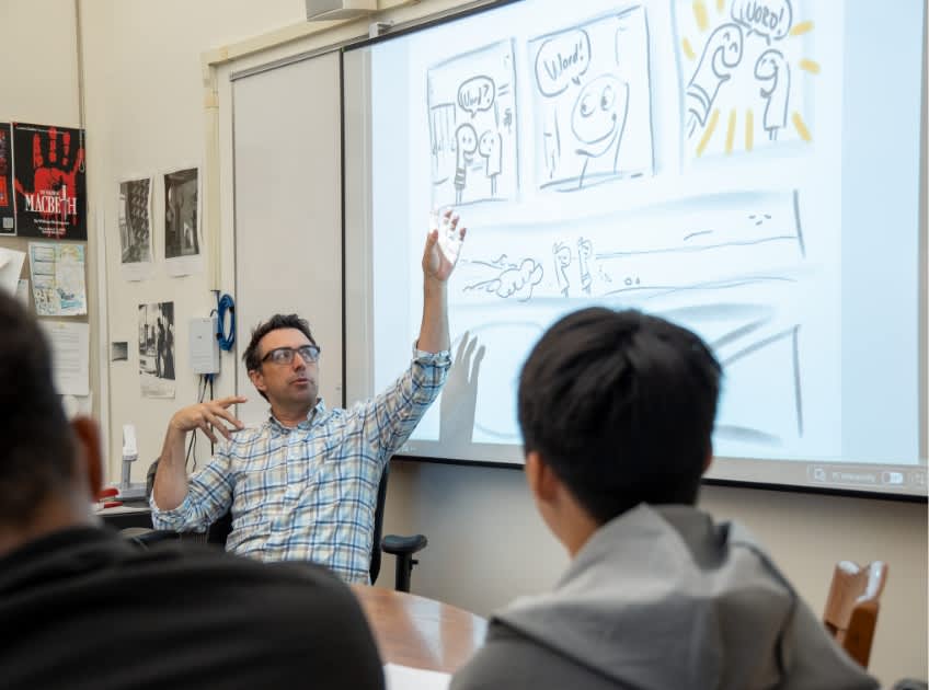 A teacher at the whiteboard with a graphic novel page projected on the screen and a student’s hand raised