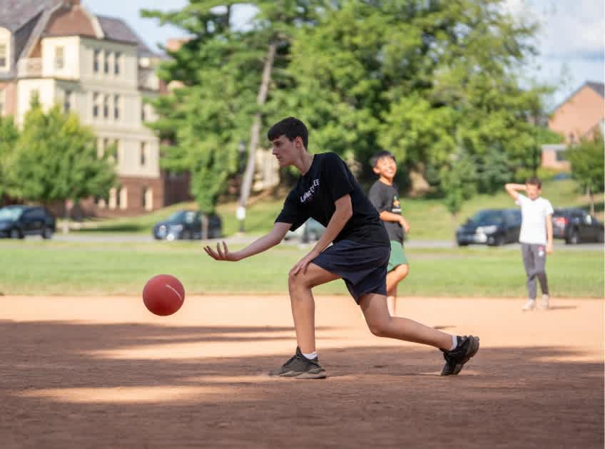 A student kicking a ball on the grass field with the campus buildings behind