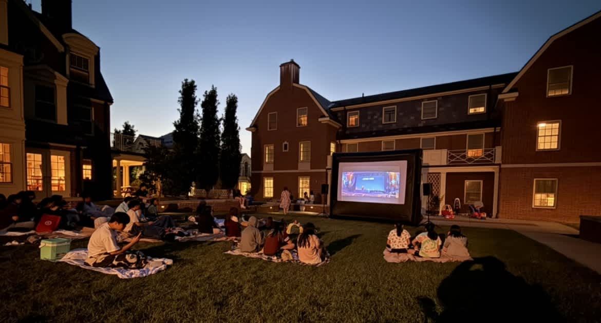 Students watching a film on a screen set up outdoors in the courtyard at dusk
