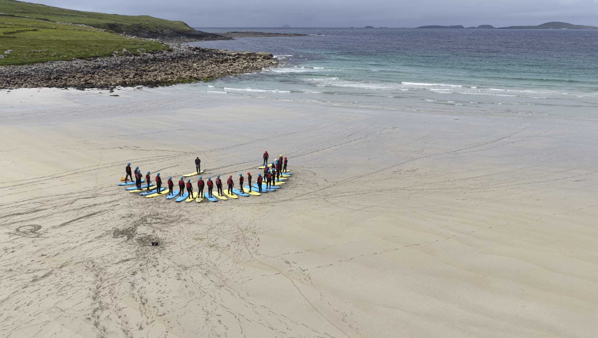 A long line of students carrying surfboards across the sand at the start of a surf lesson.