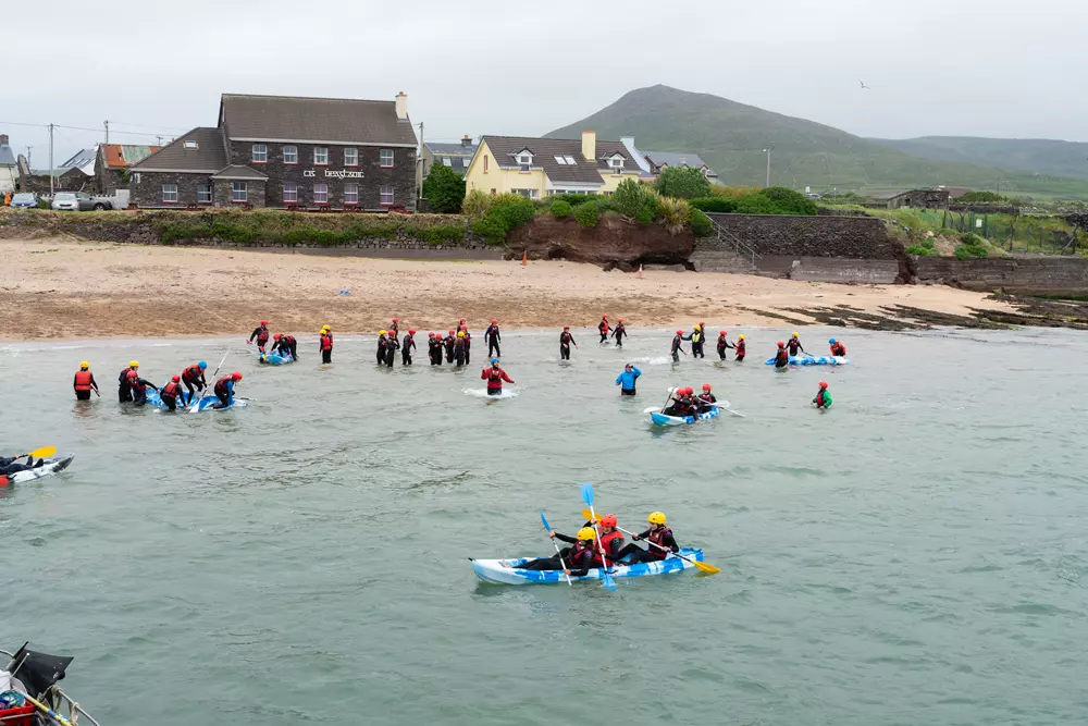 Students kayaking in shallow sea water in front of a village building on the Dingle coast