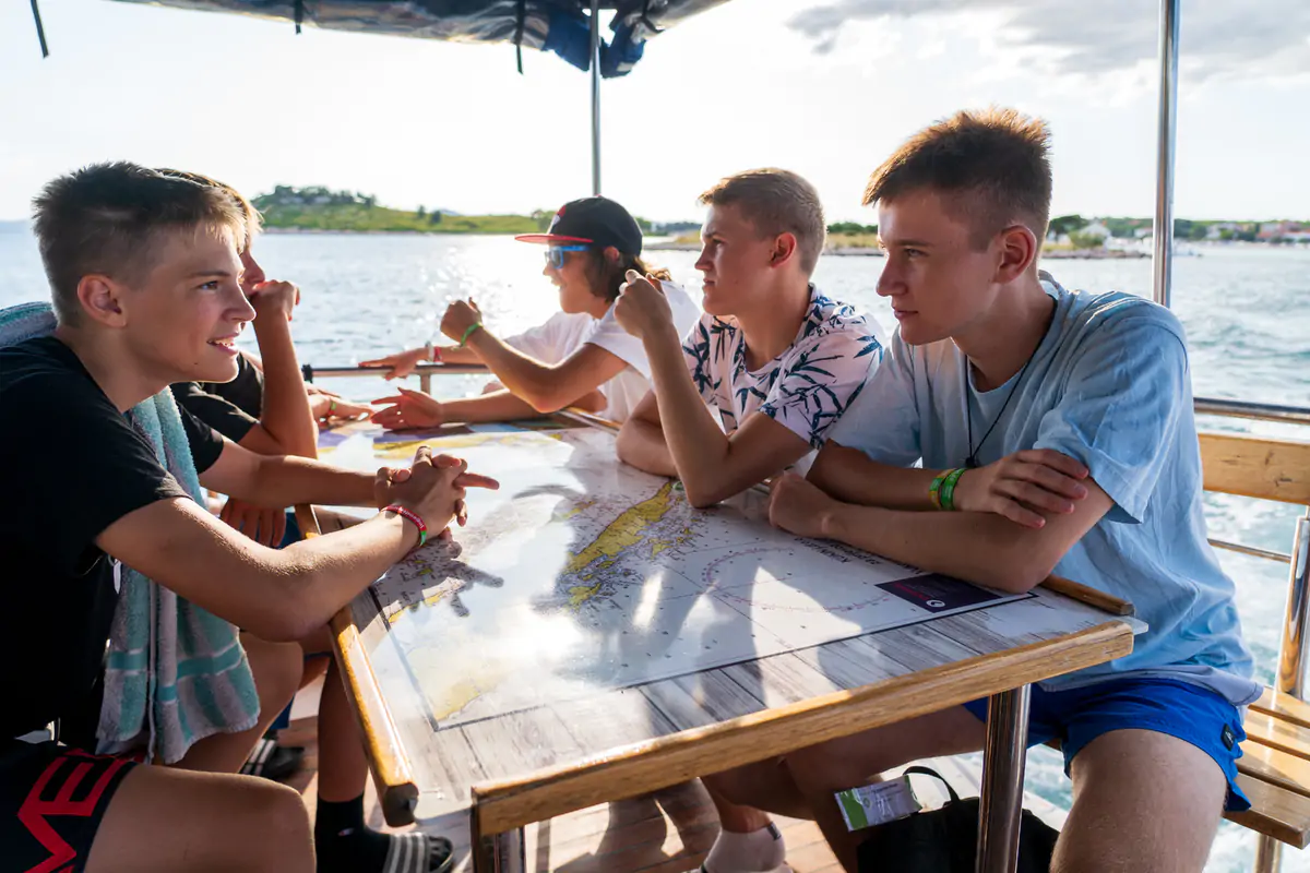 Group of teenagers playing cards around a table on a shaded terrace by the water