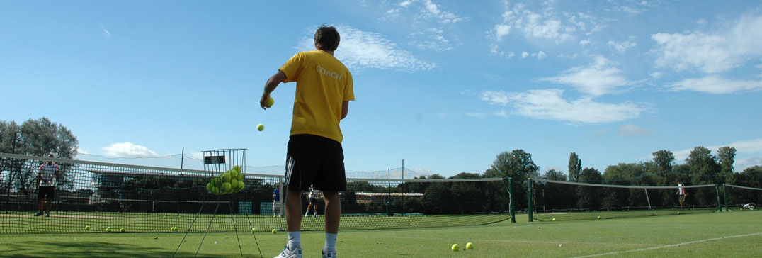Coach serving on a grass court under a blue sky at the Oxford tennis camp