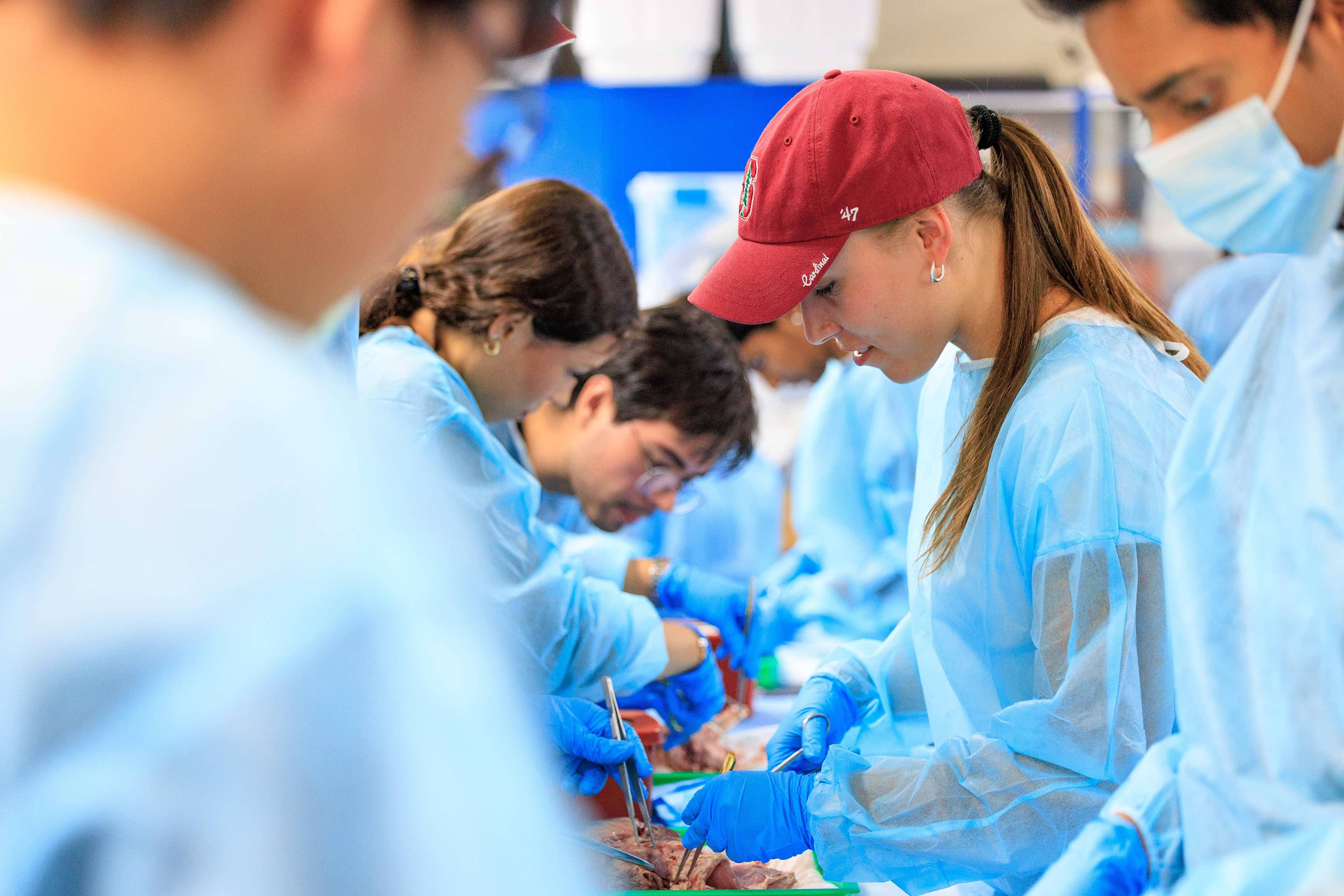 Students in blue surgical gowns, caps and gloves leaning over a table during a hands-on medicine bootcamp.