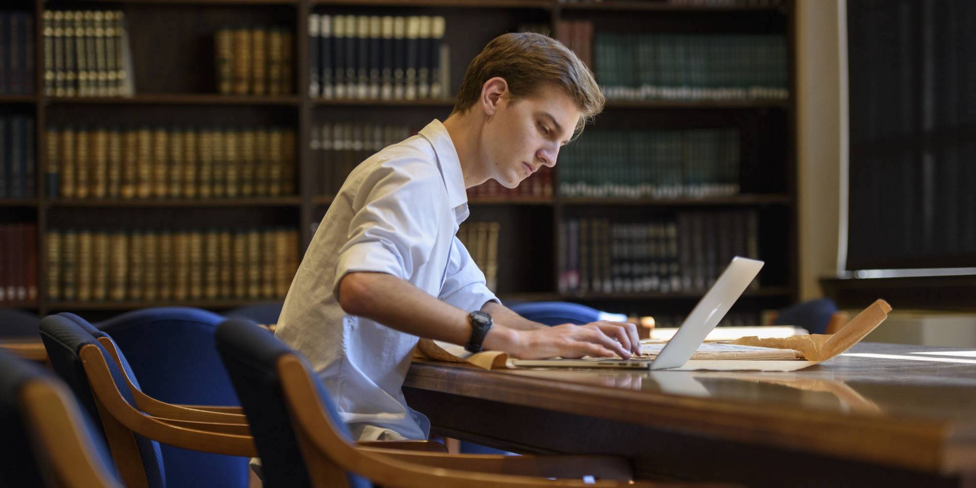 A student working with a laptop and books at a wooden desk in a wood-panelled Stanford library reading room.