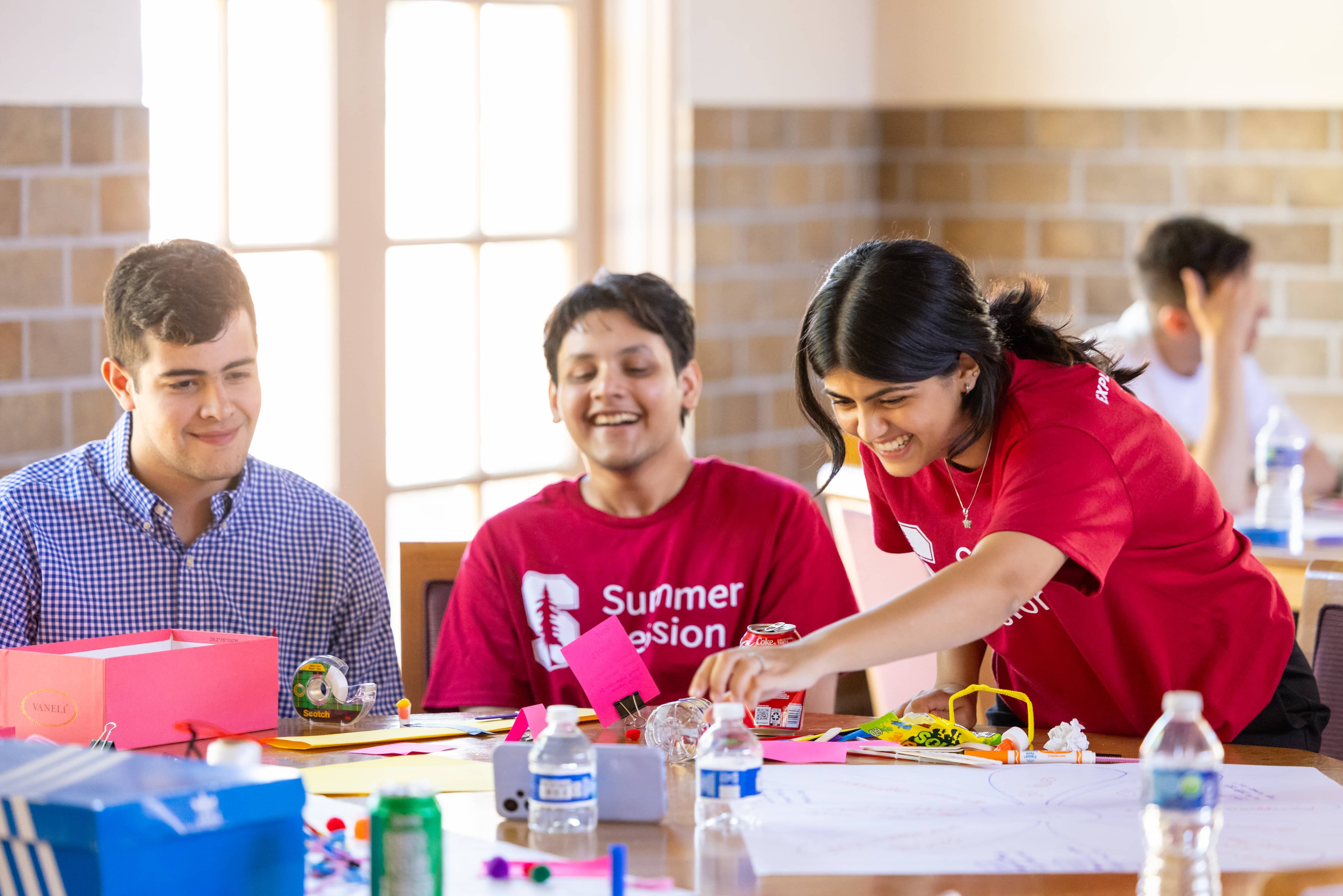 Students in Stanford Summer Session shirts working together over materials spread across a table in a sunlit hall.