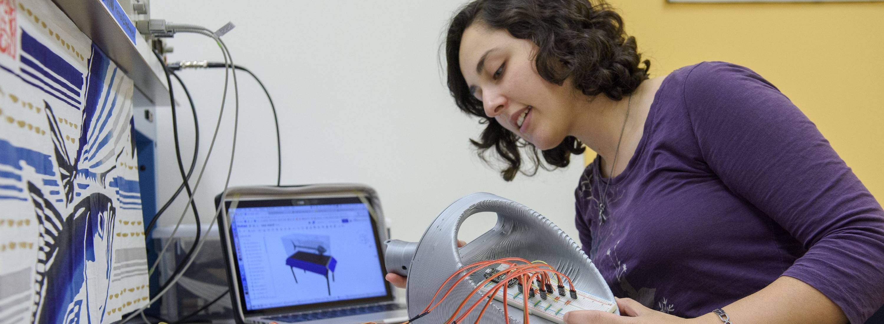 A student holding a fabricated component beside a tablet showing the digital model it was made from.