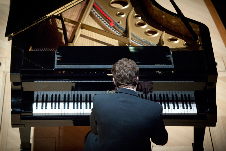 A pianist seen from behind at a grand piano on the concert hall stage