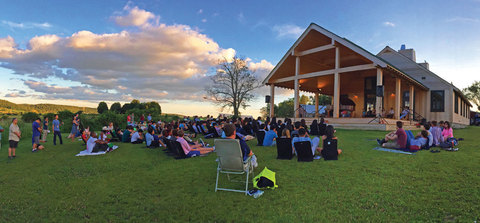 A large group of students sitting in a circle on the grass in front of the farm building at sunset