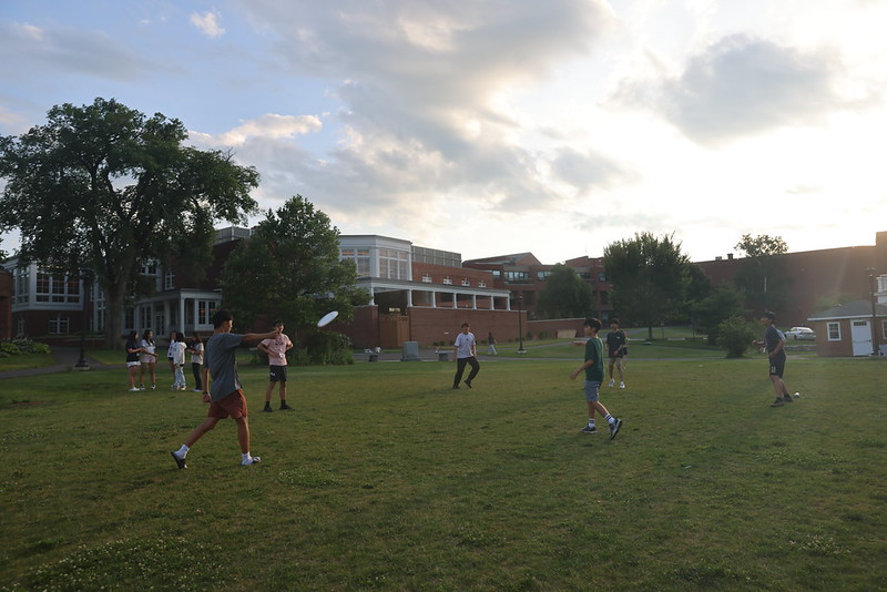 Summer Portals students throwing a disc on the lawn at dusk with the campus buildings behind them