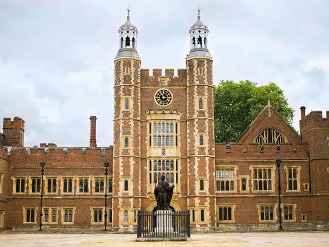 A brick tower with a clock and twin turrets on the Eton College campus, framed by trees on a clear day