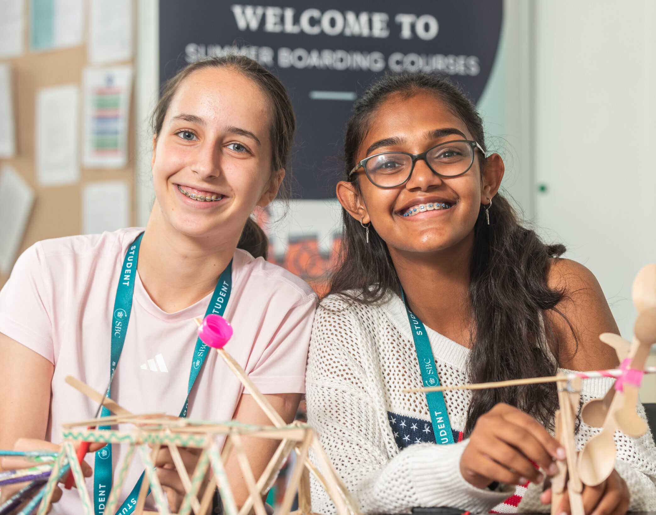 Two students smiling in a classroom as they hold up a small structure they have built from wooden sticks during a workshop