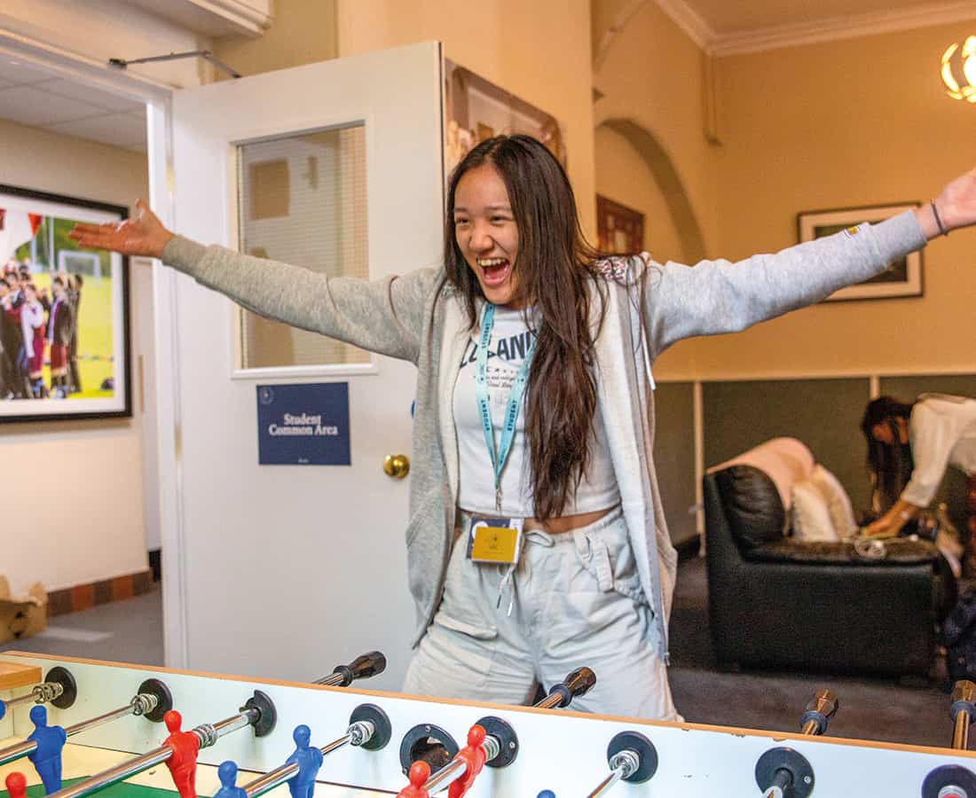 A student laughing with her arms thrown wide in the student common area, with armchairs and a noticeboard behind her
