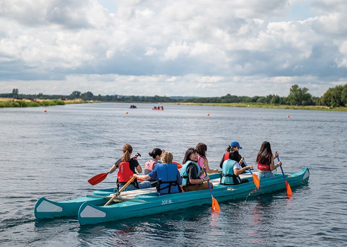 Camp students in buoyancy aids paddling two long bell boats together on open water near the Eton College campus