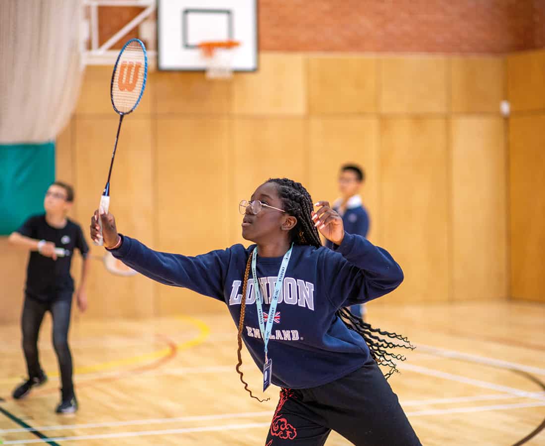 A student reaching up to play a shuttlecock during a badminton game in the wooden-floored sports hall, with other students playing behind her