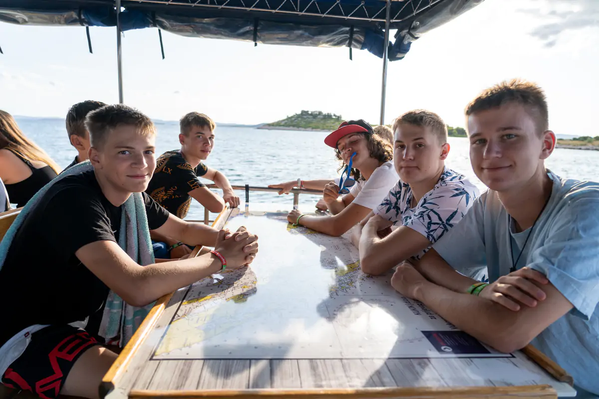 Teenagers sitting around a table on a shaded waterside terrace in Pakostane