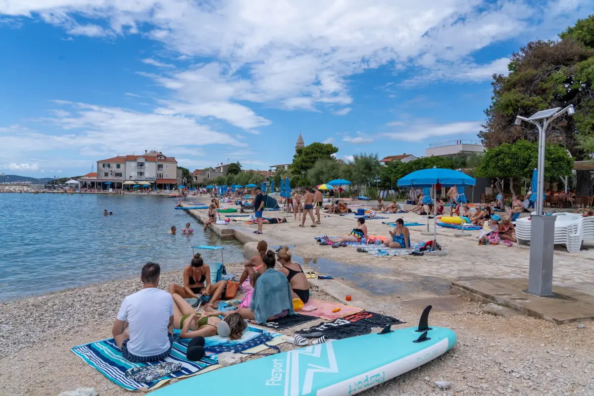 Pebble and sand beach at Pakostane with bathers, pine trees and the promenade behind