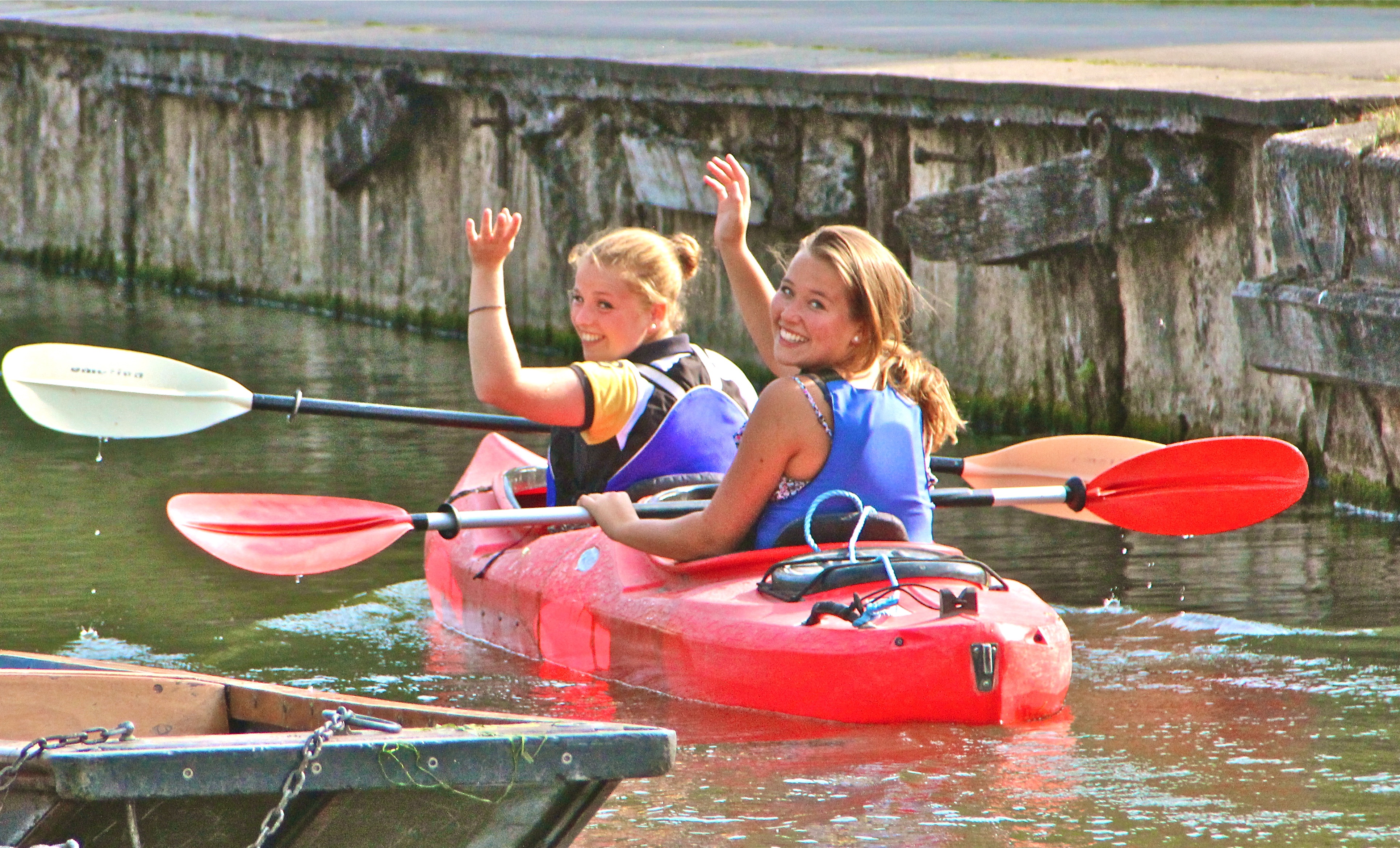 Two ISSOS students in a red two-person kayak, turning to wave with their paddles raised