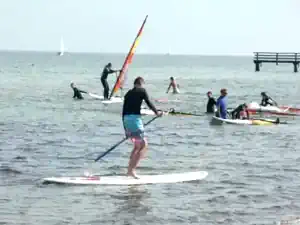 Participant stand-up paddling with windsurfers in the background at Schubystrand