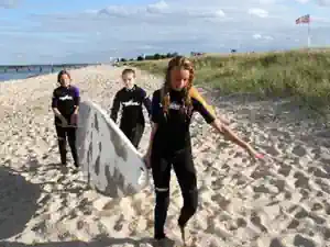 Three children in wetsuits carrying a surfboard across the beach from the surf school at Schubystrand