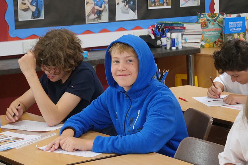 Teenagers write at desks in a classroom with a display board and stationery trays