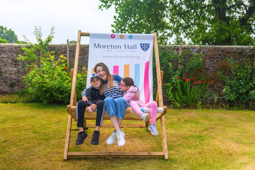 A woman and two children sit on an oversized deckchair with a Moreton Hall banner in a walled garden