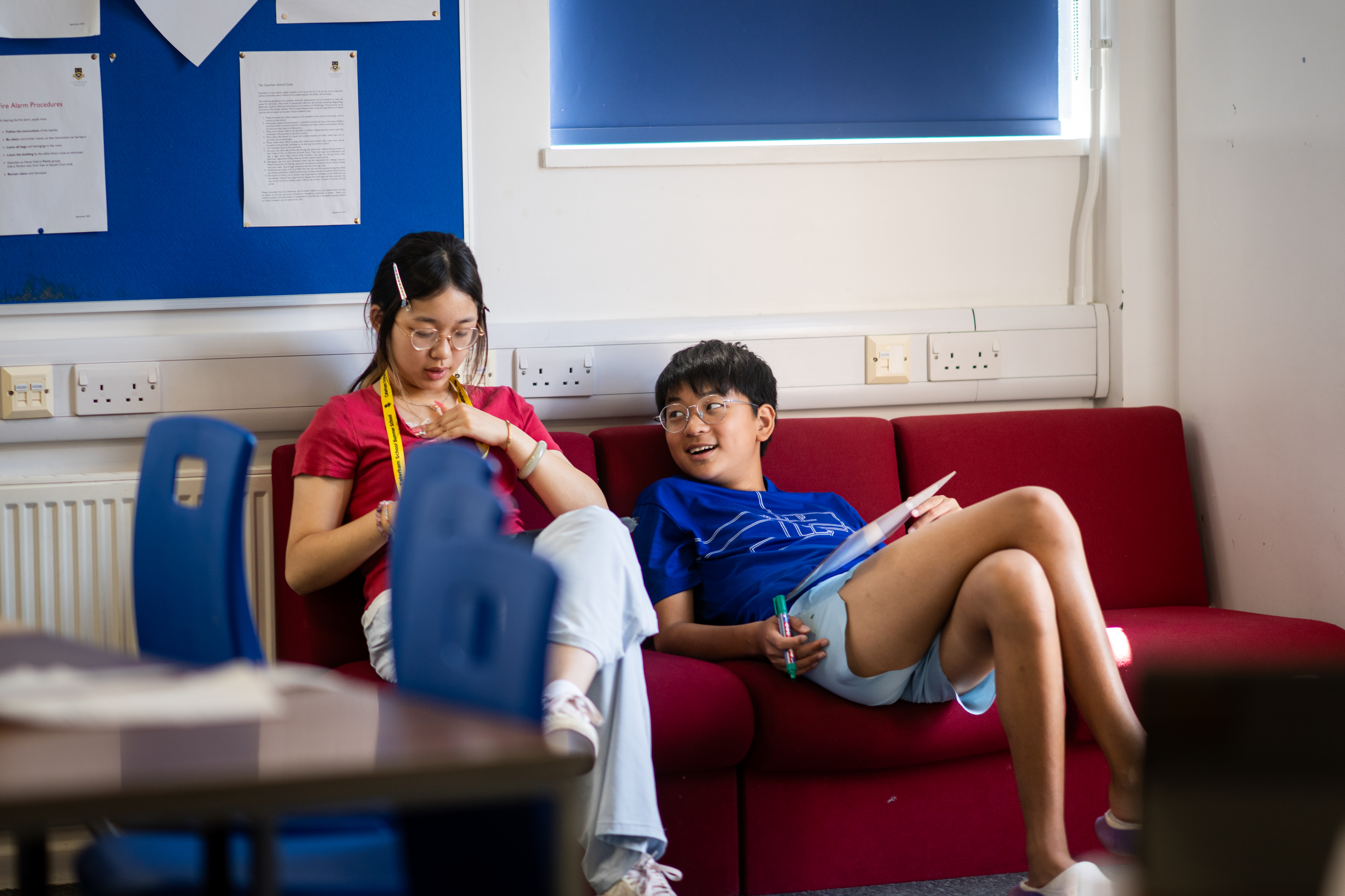 Two students sitting on a red sofa in a common room, one reading, cushions and a low table beside them