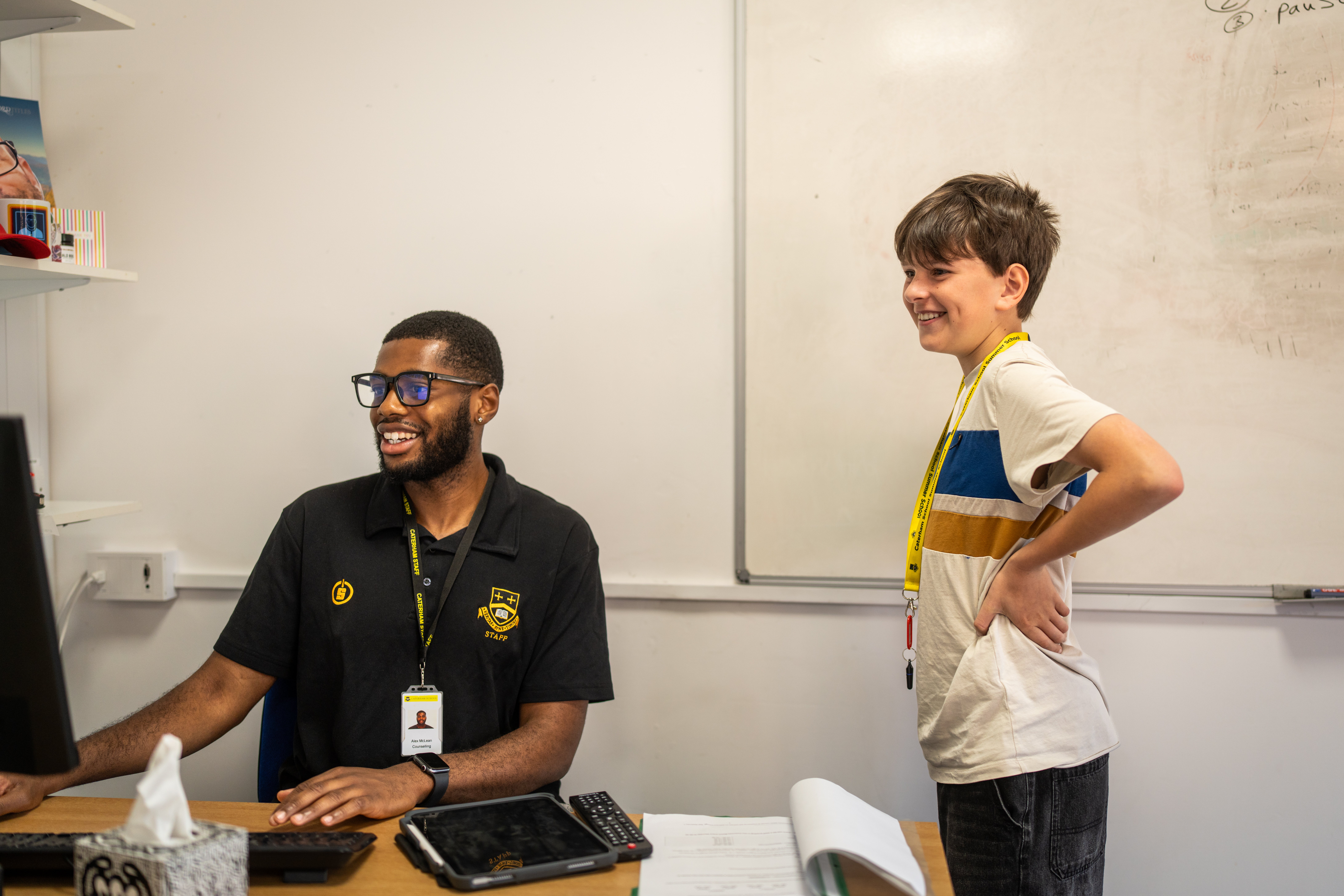 Smiling member of staff at a desk with a laptop talking to a student standing beside him in a small office
