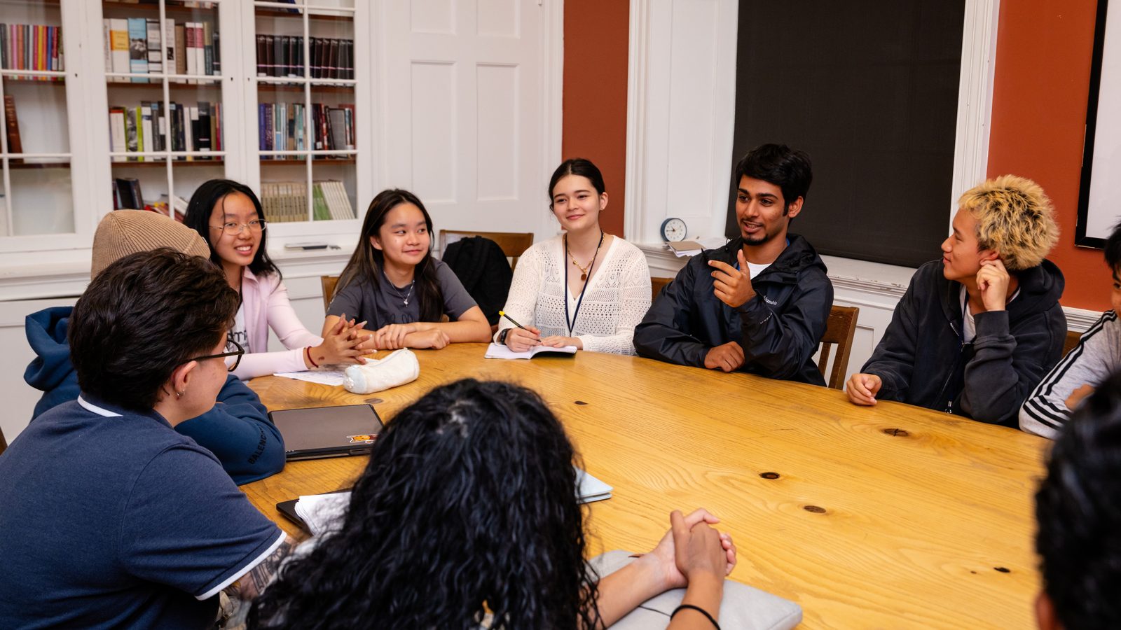 Students seated around a large wooden seminar table in a book-lined room, several with notebooks open, one speaking while the others listen.