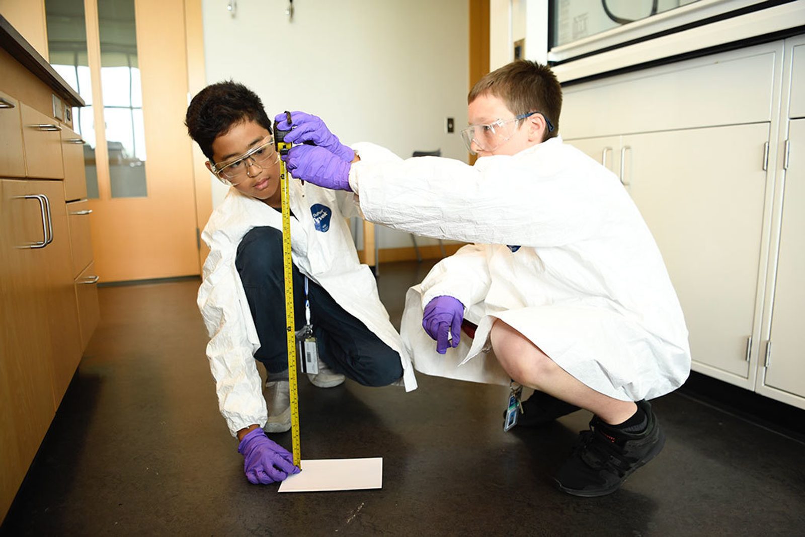 Two students in white protective coveralls, safety goggles and purple gloves crouch beside a laboratory wall, measuring a mark with a tape measure.