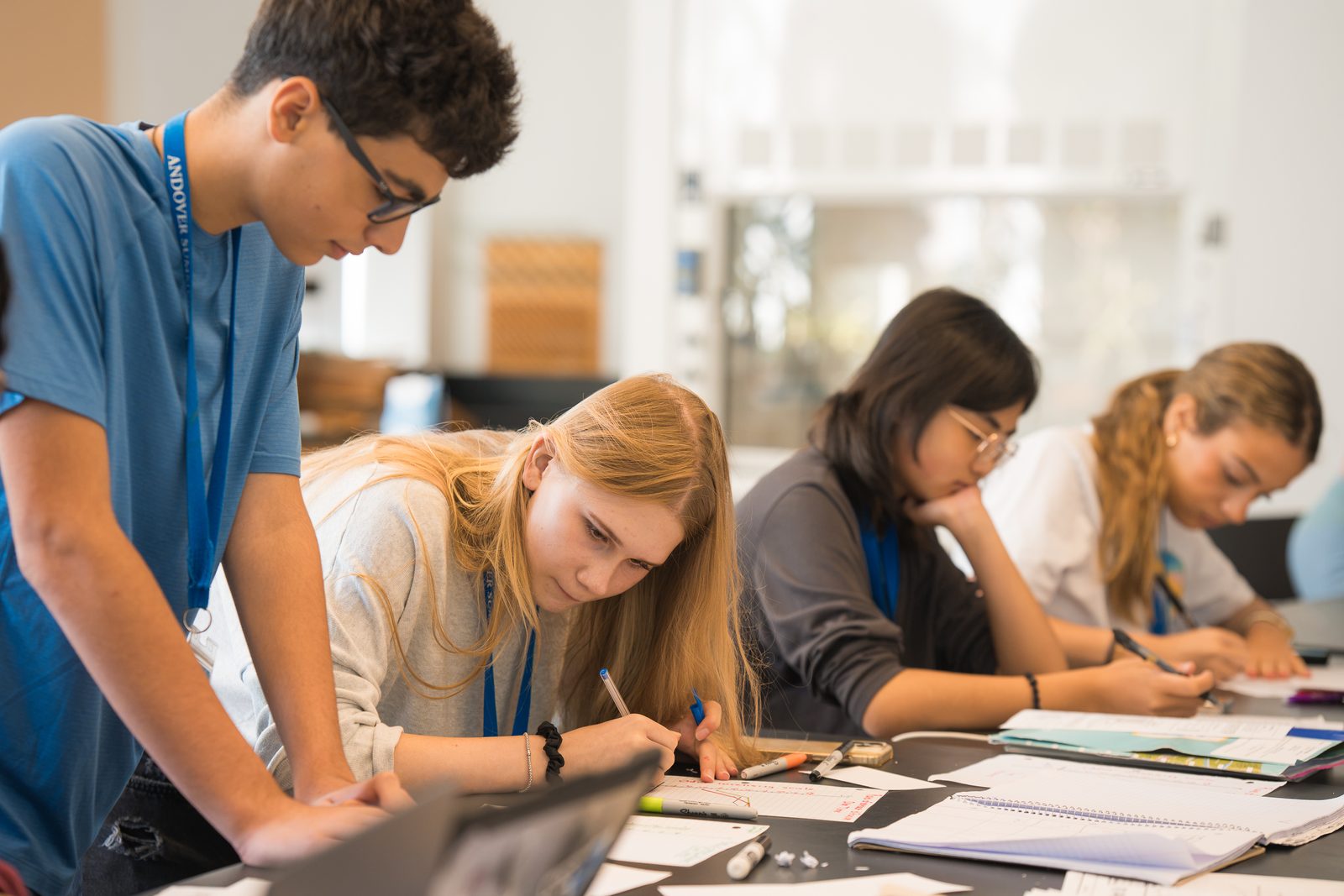 Students working in notebooks at desks in a bright classroom, wearing programme lanyards, with an instructor leaning in over the desks.