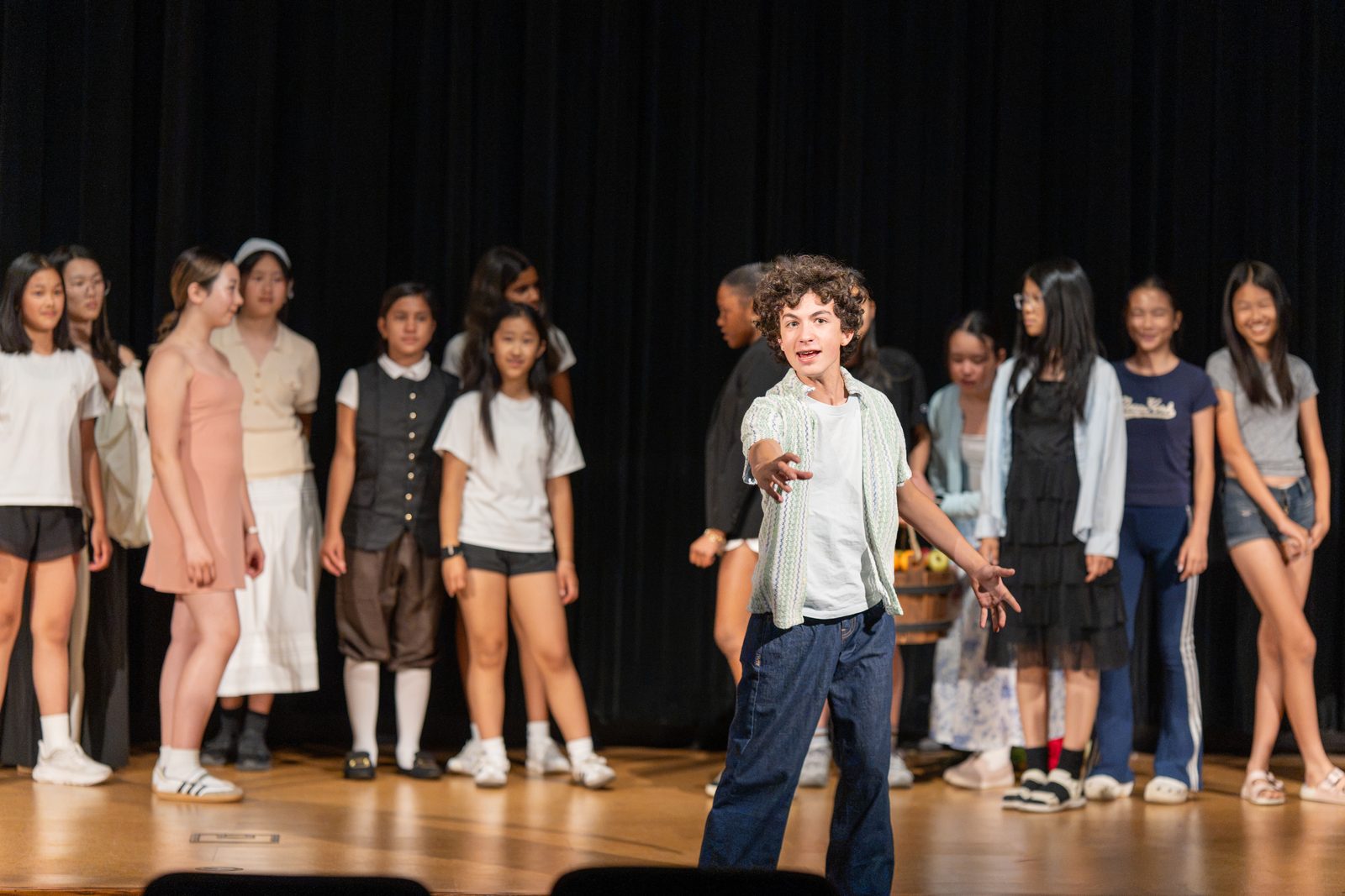 A line of students stands across a darkened stage during a performance while one student steps forward mid-gesture.