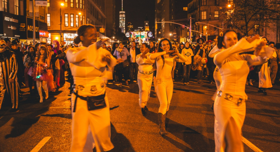 Costumed dancers in orange outfits performing in the Halloween parade on a Manhattan street at night