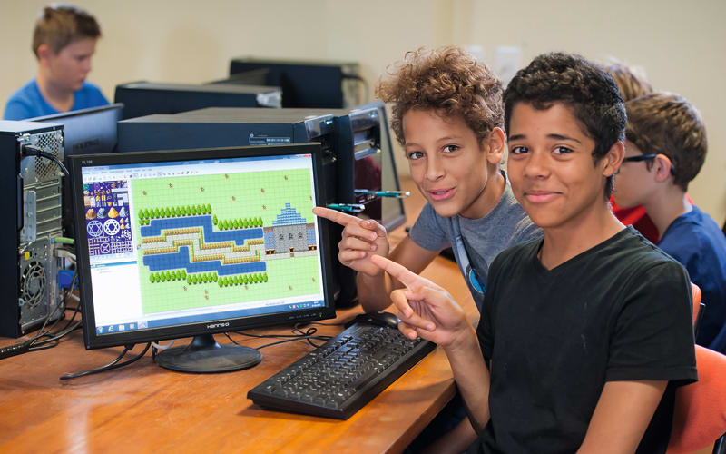Two participants at a desktop computer showing a game level they are building, with other participants working at machines behind them