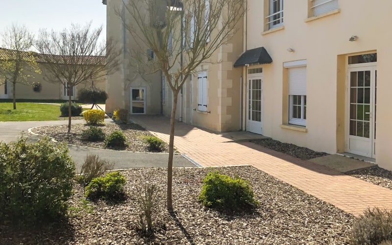 Exterior of the residence at the centre: a two-storey building with shuttered windows, a paved path and planted beds in front of it