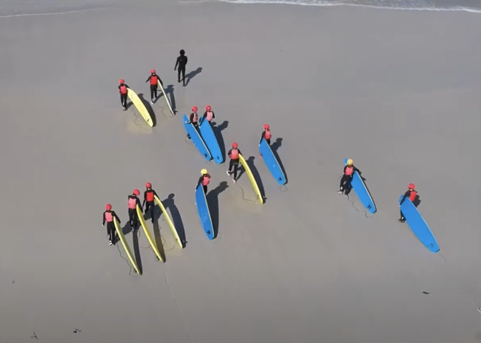 Aerial view of students carrying surfboards down the beach to the water.