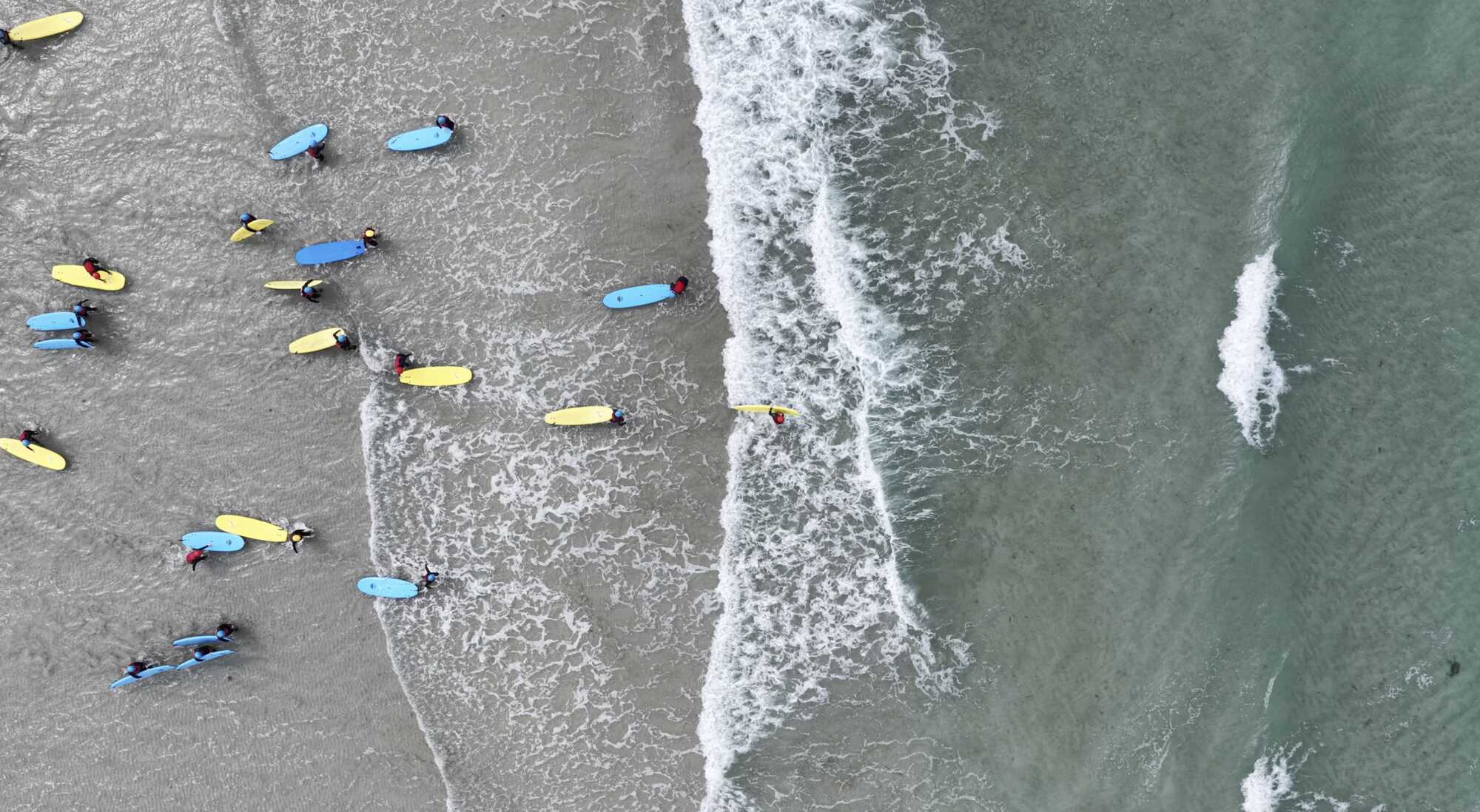 Aerial view of students with surfboards waiting for waves off the beach.