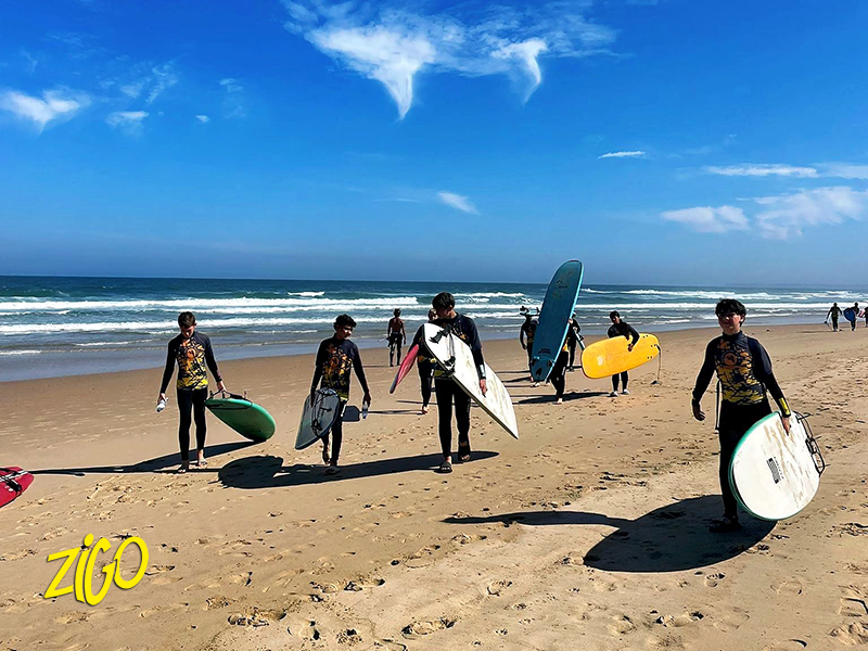 Young people in wetsuits carrying surfboards across a wide beach towards the sea